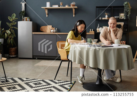 Wide shot of Asian and Caucasian female university students sitting at dining table in modern apartment preparing for finals, copy space 113255755