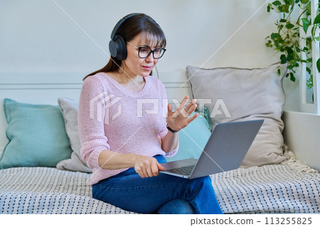 Middle aged woman in headphones having video call on laptop sitting at home 113255825
