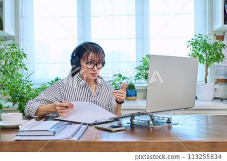 Mature woman in headphones having video conference using computer in home office 113255834