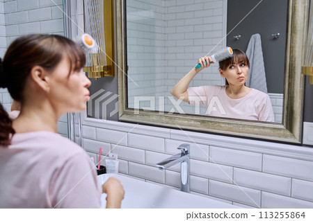 Middle-aged woman with hairspray comb looking in mirror, doing hair styling, in bathroom Middle-aged woman with hairspray comb looking in mirror, doing hair styling, in bathroom 113255864