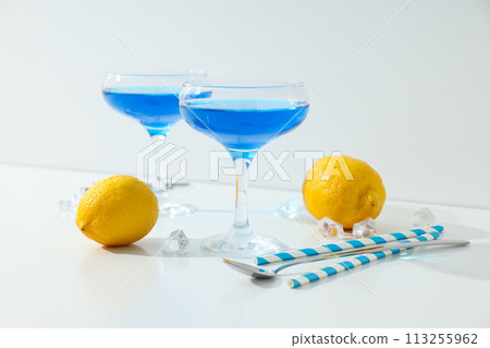 Blue cocktail in glass, whole lemon, straws and ice cubes on white background Blue cocktail in glass, whole lemon, straws and ice cubes on white background 113255962