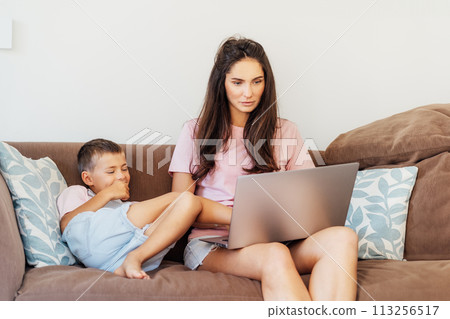 Young mother trying to work on laptop at home while her son wants to play. Childcare and working mom concept. Child make noise and disturb a woman at work. Homeschooling and freelance work. 113256517