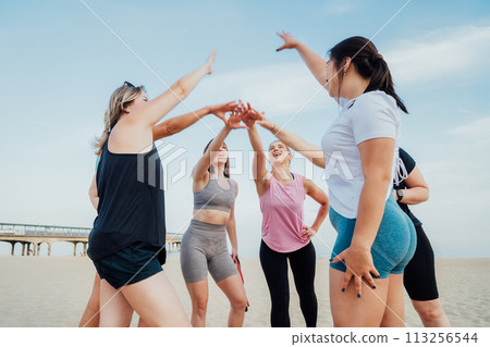 Happy fitness class giving high five after completing workout on beach. Multi aged women motivated after session together, engaged in team building, join hands for shared goal or success at training. 113256544