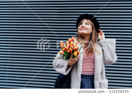 Portrait of happy, young smiling fashion woman enjoying the moment, holding bunch of fresh tulip flowers on the gray striped wall background. Urban city street fashion. Spring mood. Selective focus. 113256545