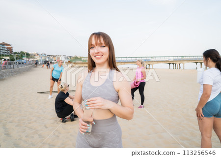 Young athletic female coach drinking water from reusable glass bottle while taking rest, refreshing during group workout class exercise on the beach. Sport for health and wellbeing. Active lifestyle. 113256546