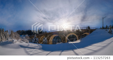 Stone viaduct (arch bridge) on railway through mountain snowy fir forest. Snow drifts  on wayside and hoarfrost on trees and electric line wires. 113257263