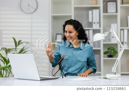 Cheerful professional woman waving and smiling during a podcast recording in a bright home office setup. Cheerful professional woman waving and smiling during a podcast recording in a bright home office setup. 113258661