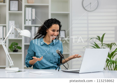 Cheerful professional woman recording a podcast in a contemporary home office set up with a laptop and microphone. 113258662