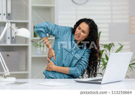 A woman in a blue shirt feels severe back pain, wincing in discomfort while sitting at her office desk with a laptop. 113258664