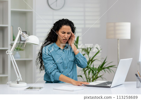 A distressed Hispanic woman is holding her ear in pain while working at her home office. She looks concerned and uncomfortable. 113258689