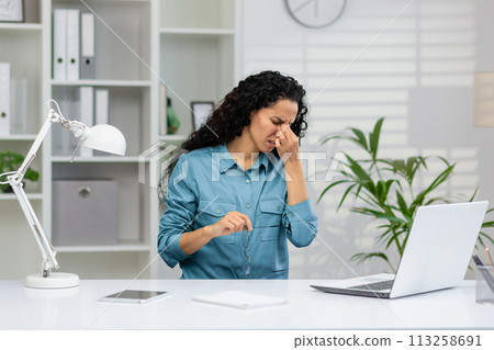 A professional businesswoman looks stressed and tired, rubbing her temples to relieve a headache while working in an office setting. 113258691