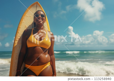Plus size African American woman wears yellow swimwear and standing with surfboard on the beach 113258940
