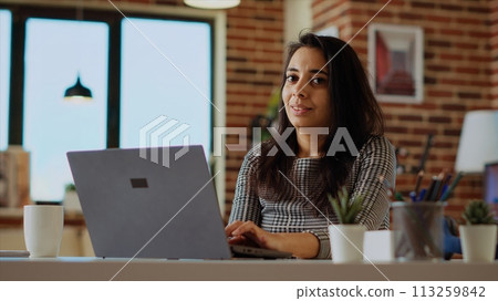 Portrait of smiling remote student typing data on keyboard and taking notes, catching up on homework. Cheerful scholar staying at home doing various tasks at clean wooden desk, camera B 113259842