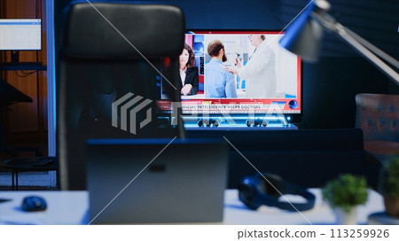 Smart TV left open on news broadcast in empty home office with desk filled with business items. Television set showing VOD channel with Laptop, headphones, plants in blurry foreground, camera A 113259926
