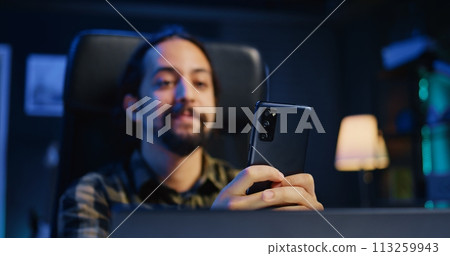 Happy man at home texting friend on cellphone, taking break from remote job in personal office. Joyful freelancer relaxing in apartment, talking with mate on internet using phone, camera B 113259943