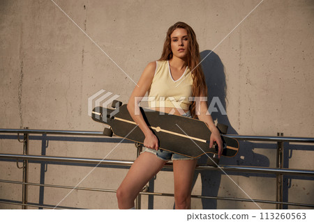 Redheaded sporty woman posing for camera with her skateboard over concrete wall Redheaded sporty woman posing for camera with her skateboard over concrete wall 113260563