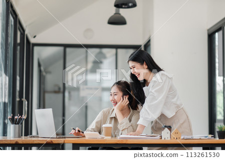 Two joyful colleagues share a light-hearted moment, with one seated at a laptop and the other standing beside her, in a spacious and well-lit office. 113261530