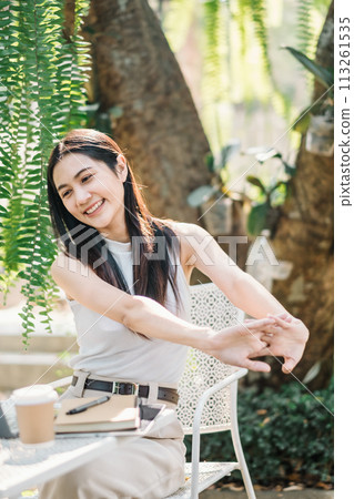 Relaxed female freelancer stretching arms while taking a break from work on her laptop in a lush garden cafe setting. Relaxed female freelancer stretching arms while taking a break from work on her laptop in a lush garden cafe setting. 113261535