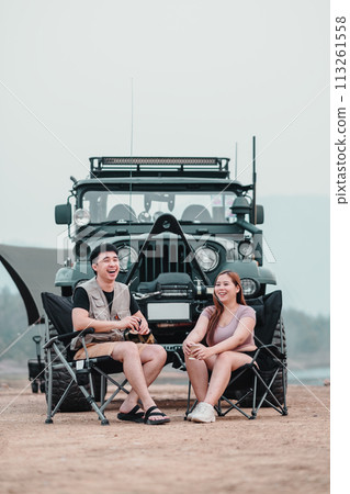 Man and woman laugh joyfully while sitting in camping chairs, with a off-road car in the backdrop, enjoying their outdoor adventure. 113261558