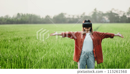 Young woman stands with arms wide open, wearing a VR headset in a lush green field, embracing the virtual experience. 113261568