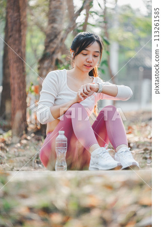 Smiling woman in sportswear checking her fitness tracker after exercising, seated in a serene forest area. 113261585