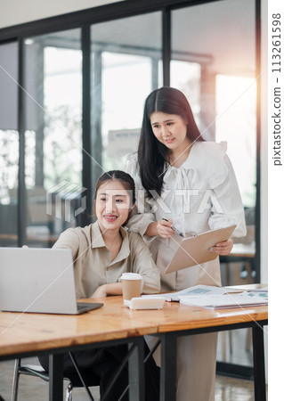 Two smiling female colleagues collaboratively working on a project with a laptop and coffee in a bright office space. 113261598