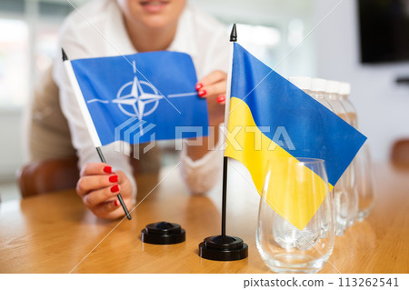 Flags of NATO and Ukraine in hands of woman preparing meeting room for negotiation Flags of NATO and Ukraine in hands of woman preparing meeting room for negotiation 113262541