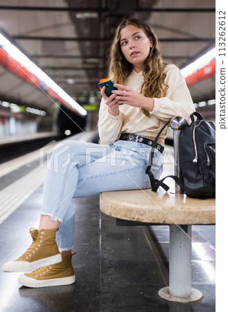 Portrait of a pensive girl sitting on a subway platform bench with a mobile phone Portrait of a pensive girl sitting on a subway platform bench with a mobile phone 113262612