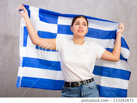 Sad young woman holding Greece flag against unicoloured background Sad young woman holding Greece flag against unicoloured background 113262672