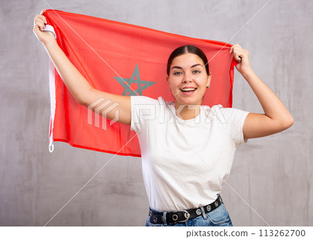 Happy young woman posing and smiling, standing with flag of Morocco against gray wall 113262700