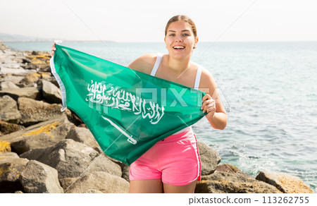 Portrait of positive young woman with the flag of Saudi Arabia on the beach on sunny day 113262755