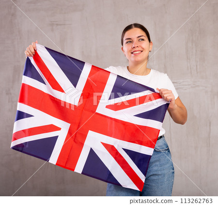 Satisfied young woman holding national flag of Great Britain on gray wall background Satisfied young woman holding national flag of Great Britain on gray wall background 113262763