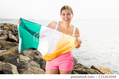 Young woman on coast posing with flag of Ireland Young woman on coast posing with flag of Ireland 113263040