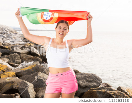 Enjoying vacation in Portugal. Young woman with national Portuguese flag on the seashore on sunny day Enjoying vacation in Portugal. Young woman with national Portuguese flag on the seashore on sunny day 113263118