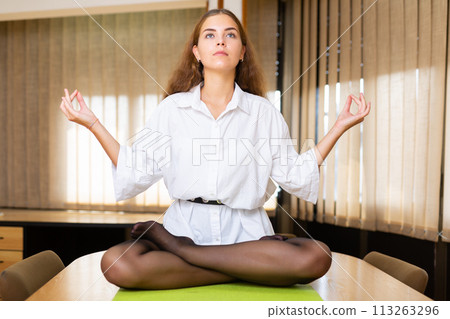 Relaxed young woman meditating in Padmasana pose on table in office 113263296