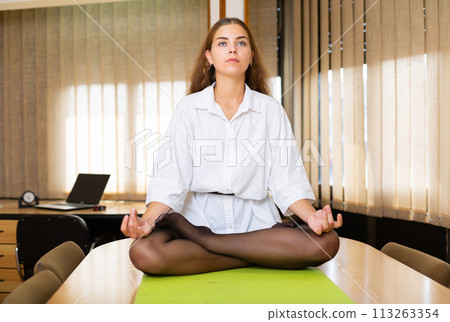 Relaxed young woman meditating in Padmasana pose on table in office Relaxed young woman meditating in Padmasana pose on table in office 113263354