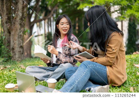 Two young Asian female students are sitting on the grass in a park, studying together. 113265244