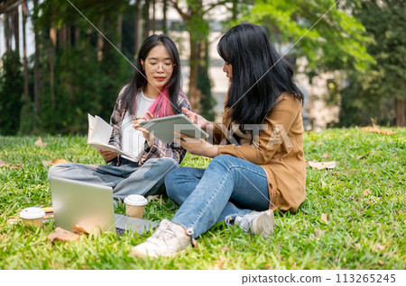 Two young Asian female students are sitting on the grass in a park, studying together. 113265245