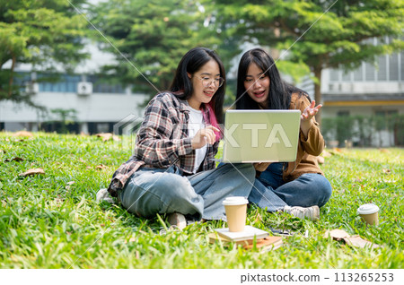 Two young Asian female college students are doing homework on a laptop together, sitting on grass. 113265253