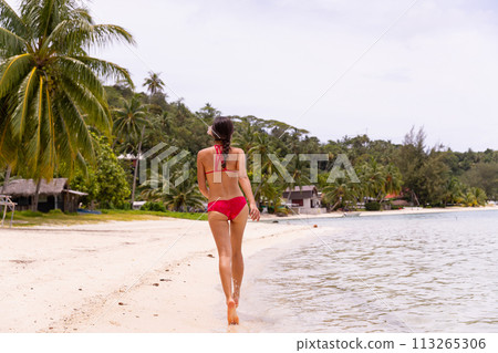 Beach Stroll Serenity: Woman in Red Bikini Walking on Matira Beach, Bora Bora, Tahiti, French Polynesia. This image is completely unretouched and model is with no makeup. Raw Image 113265306
