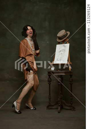 Eccentric, emotional young man dressed as woman in old-fashion silky dress posing near self-portrait on easel against vintage background. 113269124