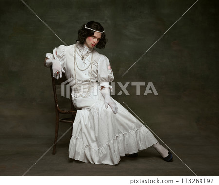 Pensive man wearing period gown and wig, adorned with pearls and seated on wooden chair against vintage studio background. Pensive man wearing period gown and wig, adorned with pearls and seated on wooden chair against vintage studio background. 113269192
