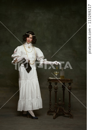 Man in white old-fashion blouse and skirt, pouring drink, adorned with pearls and beaded purse against vintage studio background. Man in white old-fashion blouse and skirt, pouring drink, adorned with pearls and beaded purse against vintage studio background. 113269217