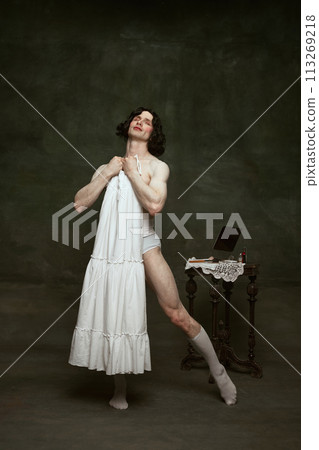 Man with flowing hair exudes confidence in white sleeveless dress, near antique makeup table at vintage room. Breaking traditions. Man with flowing hair exudes confidence in white sleeveless dress, near antique makeup table at vintage room. Breaking traditions. 113269218