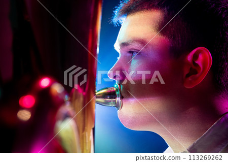 Close up side view photo of talented man, artistic musician seated, playing trumpet with soft blue-pink stage lights. 113269262