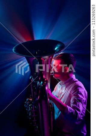 Side view photo of talented man, artistic musician seated, playing trumpet with soft blue-pink stage lights behind him. 113269263