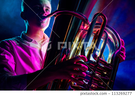 Detailed photo of tuba being played by artistic musician, highlighted by glow of dramatic stage lighting. Art. 113269277