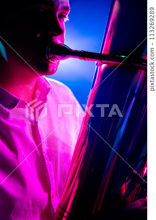 Portrait of talented musician seated, playing trumpet with soft blue-pink stage lights. Classical symphony. 113269289