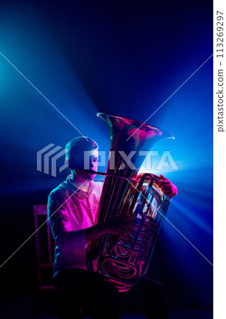 Portrait of talented musician seated, playing trumpet with soft blue-pink stage lights highlighted his silhouette. 113269297