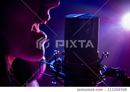 Cropped close up of female vocalist singing passionately near studio microphone with purple flare lighting. 113269306